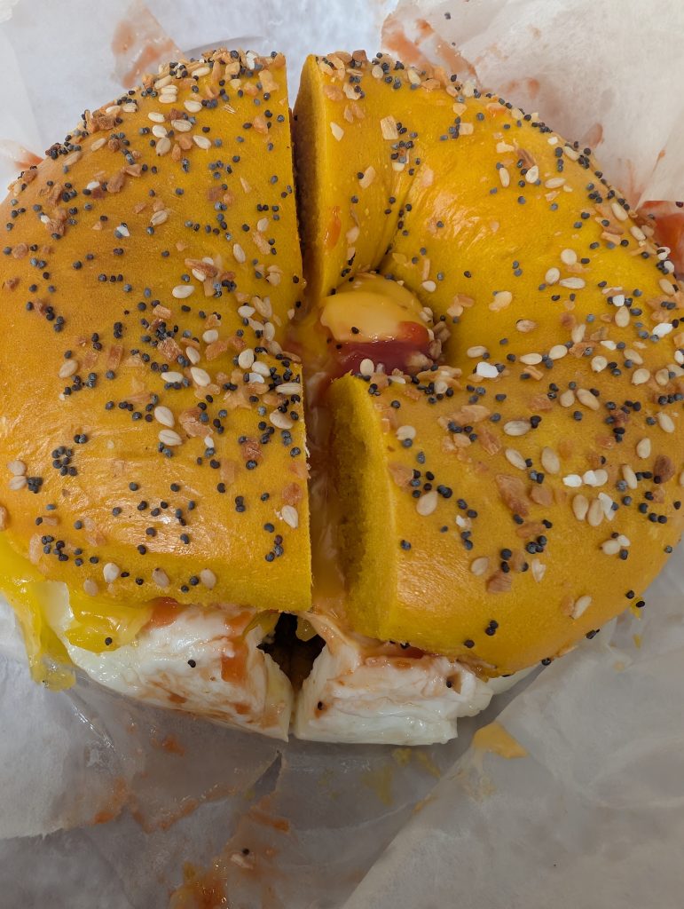 Egg everything bagel with bacon, egg, cheese, salt, pepper and ketchup on white deli paper.
