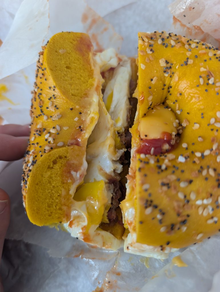 Egg everything bagel with bacon, egg, oozing cheese, salt, pepper and ketchup on white deli paper.