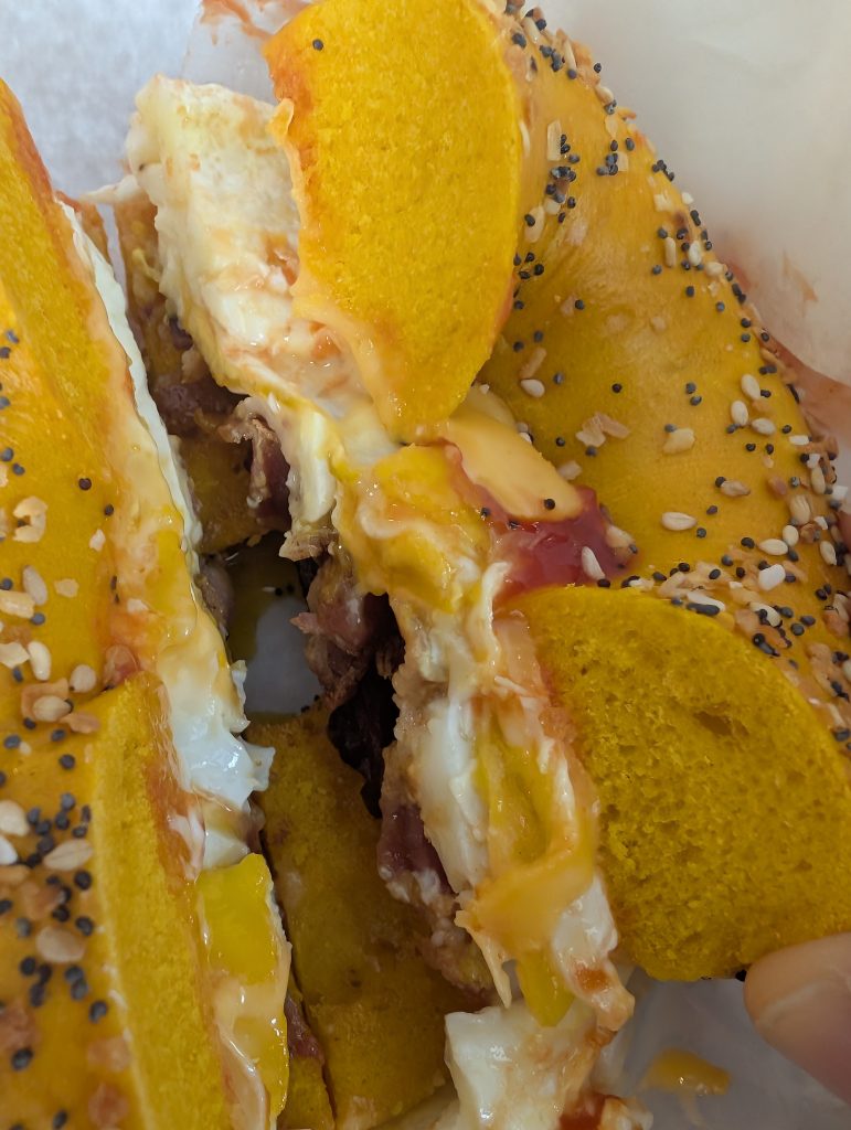 Egg everything bagel with bacon, egg, oozing cheese, salt, pepper and ketchup on white deli paper.