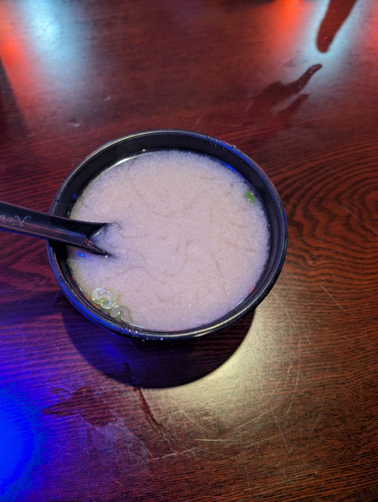 A black bowl of traditional Japanese miso soup with green onions and a spoon on a wooden table at Kira Restaurant.